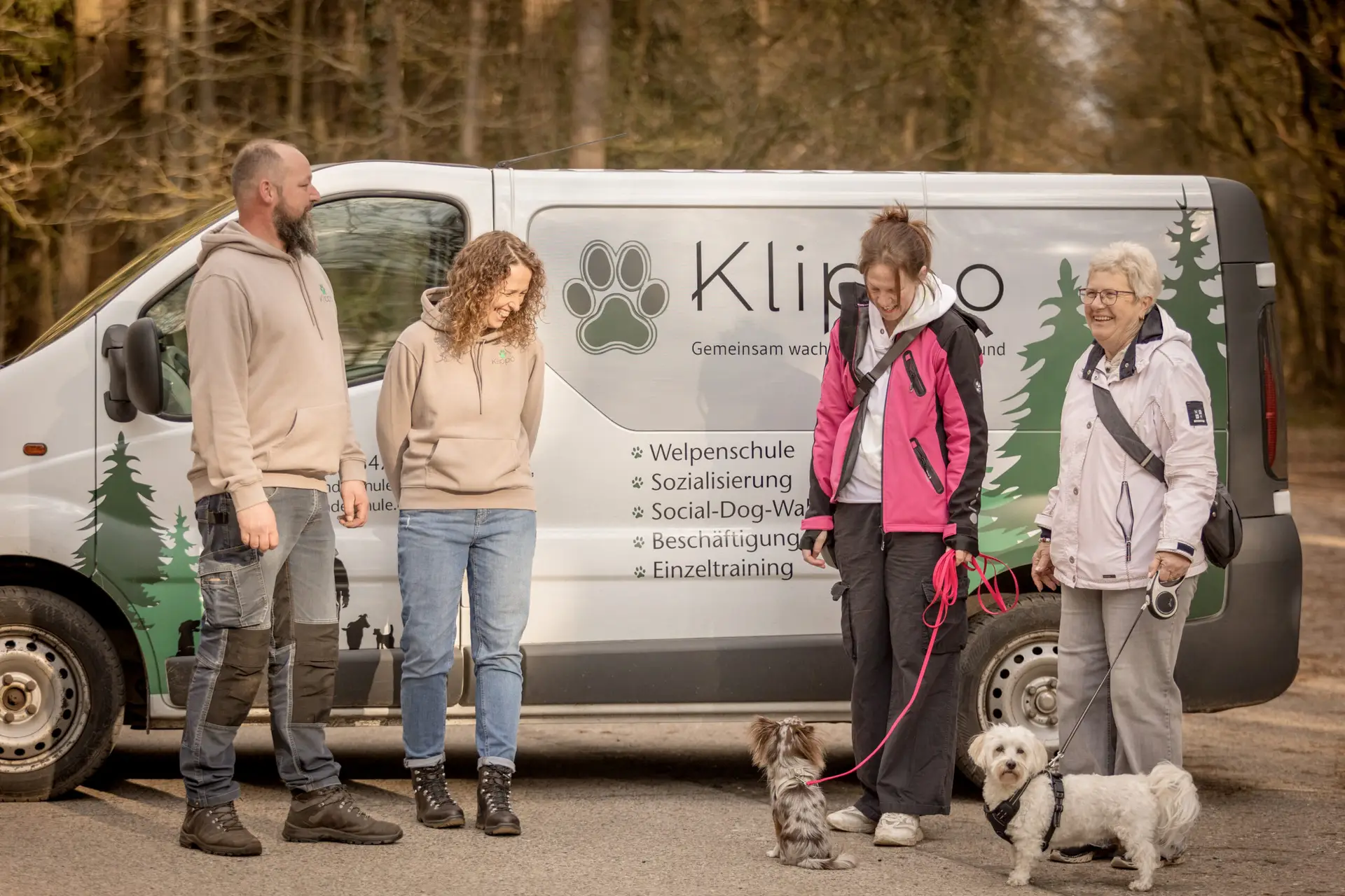 Meik und Sandra von der Hundeschule Klippo stehen mit zwei Hundebesitzern und ihren Hunden vor dem Firmenfahrzeug und posieren für ein Foto